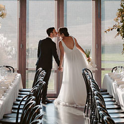 Bride and groom standing by the huge windowed wall of The Stables reception center, surrounded by floral table decor.